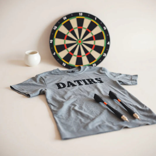 Darts-themed t-shirt laid flat with darts and a blurred dartboard in the background